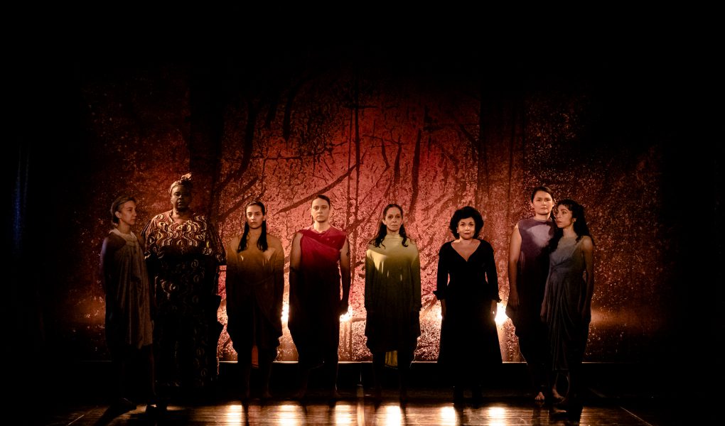 Eight women stand in front of a red backdrop. Stage photo.