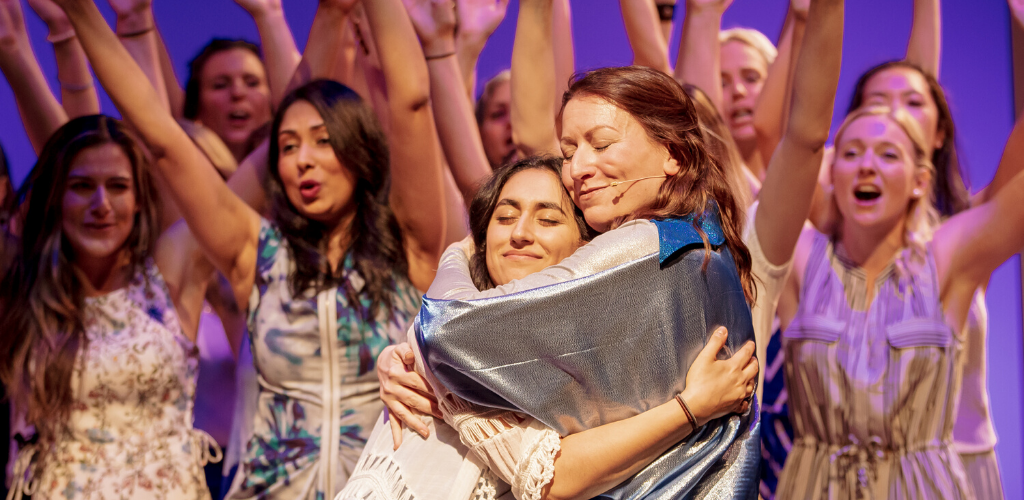 Two women hug in the foreground, a chorus of women dances in the background.