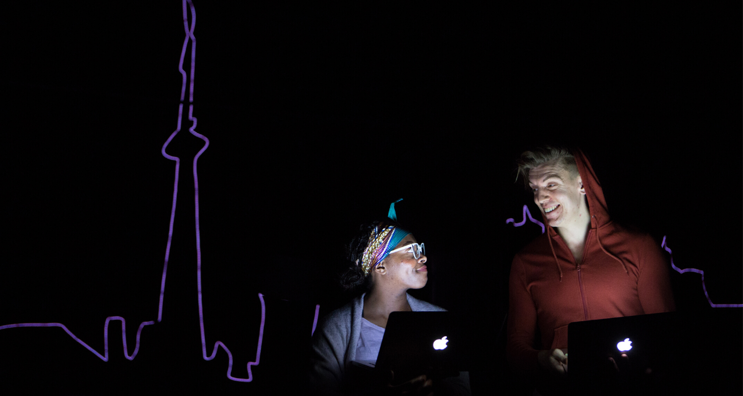 Monice Peter and Adrian Shepherd-Gawinski looking at each other under the glow of their laptops, with the outline of the Toronto skyline in the background.