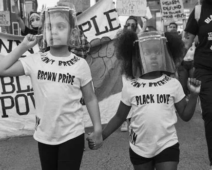Black and white image of two children, holding hands and wearing face shields, at a protest. One wears a shirt that says "Brown pride" and the other wears a shirt that says "Black love."