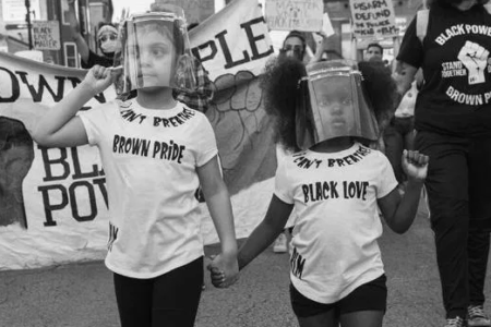 Black and white image of two children, holding hands and wearing face shields, at a protest. One wears a shirt that says "Brown pride" and the other wears a shirt that says "Black love."