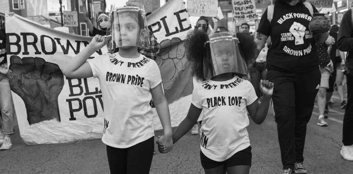 Black and white image of two children, holding hands and wearing face shields, at a protest. One wears a shirt that says "Brown pride" and the other wears a shirt that says "Black love."