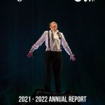 Teiya Kasahara wearing a white button down shirt tucked into black pants with black suspenders and an undone bowtie. They stand with their arms at their sides, fingers splayed, looking up joyously. Text reads: Nightwood Theatre, 2021-2022 Annual Report.