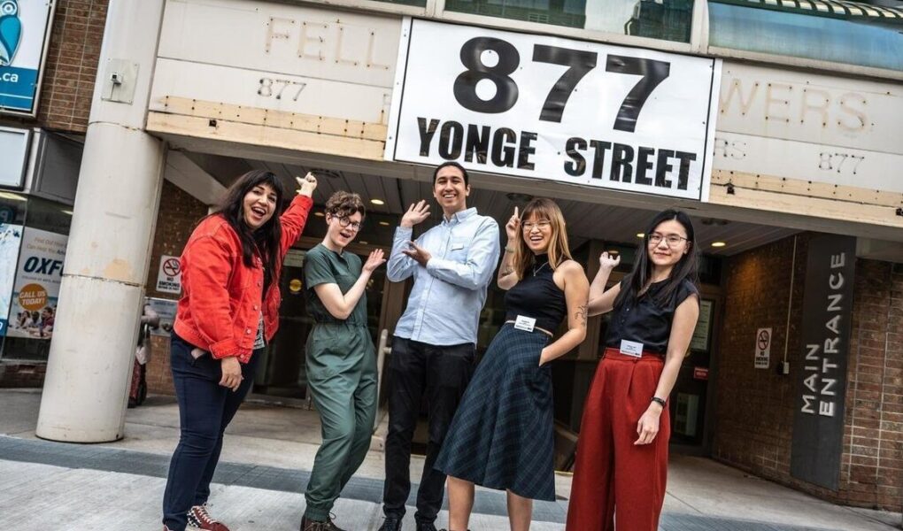 Photo of five smiling staff members in colourful upscale clothes. They pose in front of a brick and concrete building that says 877 Yonge street