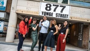 Photo of five smiling staff members in colourful upscale clothes. They pose in front of a brick and concrete building that says 877 Yonge street