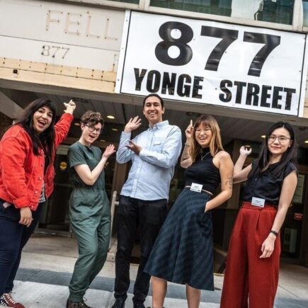 Photo of five smiling staff members in colourful upscale clothes. They pose in front of a brick and concrete building that says 877 Yonge street