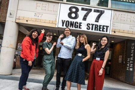 Photo of five smiling staff members in colourful upscale clothes. They pose in front of a brick and concrete building that says 877 Yonge street