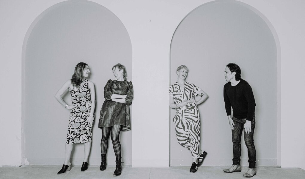 Black and white photo of Naz Afsahi, Andrea Donaldson, Jaime Martino, and Michael Mori leaning against a wall with tall arches and smiling at each other.