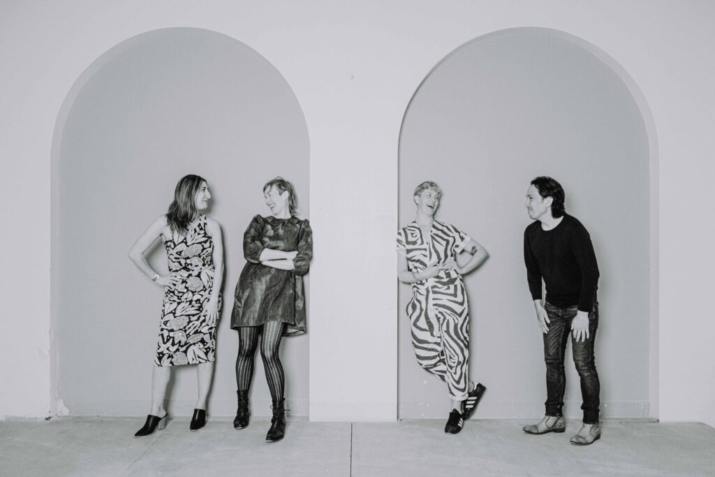 Black and white photo of Naz Afsahi, Andrea Donaldson, Jaime Martino, and Michael Mori leaning against a wall with tall arches and smiling at each other.