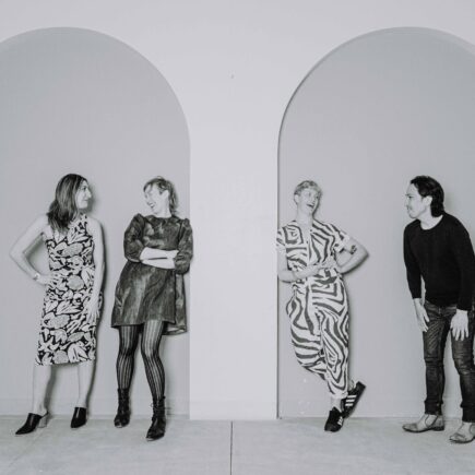 Black and white photo of Naz Afsahi, Andrea Donaldson, Jaime Martino, and Michael Mori leaning against a wall with tall arches and smiling at each other.