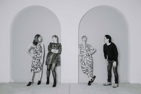 Black and white photo of Naz Afsahi, Andrea Donaldson, Jaime Martino, and Michael Mori leaning against a wall with tall arches and smiling at each other.