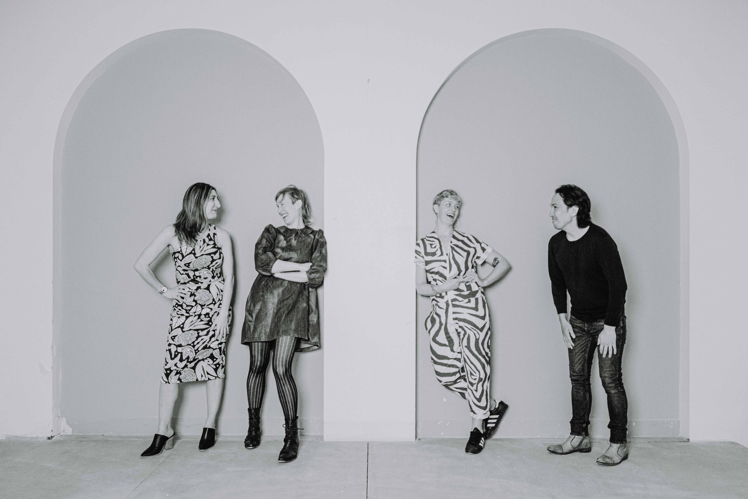 Black and white photo of Naz Afsahi, Andrea Donaldson, Jaime Martino, and Michael Mori leaning against a wall with tall arches and smiling at each other.