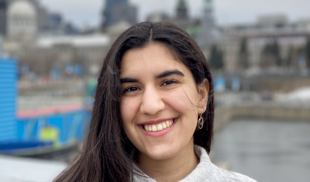 A light brown-skinned woman of Iranian descent in her early 20s, with the background of Montreal, wearing a grey and beige patterned sweater. Long, dark brown hair is parted to one side and tucked behind her ear. She has dark brown eyes and is smiling, looking at the camera.