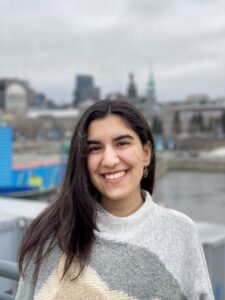 A light brown-skinned woman of Iranian descent in her early 20s, with the background of Montreal, wearing a grey and beige patterned sweater. Long, dark brown hair is parted to one side and tucked behind her ear. She has dark brown eyes and is smiling, looking at the camera.