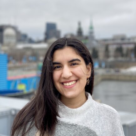 A light brown-skinned woman of Iranian descent in her early 20s, with the background of Montreal, wearing a grey and beige patterned sweater. Long, dark brown hair is parted to one side and tucked behind her ear. She has dark brown eyes and is smiling, looking at the camera.
