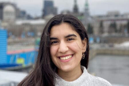 A light brown-skinned woman of Iranian descent in her early 20s, with the background of Montreal, wearing a grey and beige patterned sweater. Long, dark brown hair is parted to one side and tucked behind her ear. She has dark brown eyes and is smiling, looking at the camera.