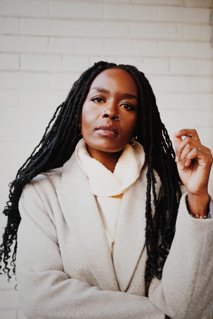 Germaine Konji, a Kenyan-Canadian with dark skin and long black faux-locks gazes at the camera. They are wearing a white turtleneck and a grey wool coat. The background is a white brick wall.
