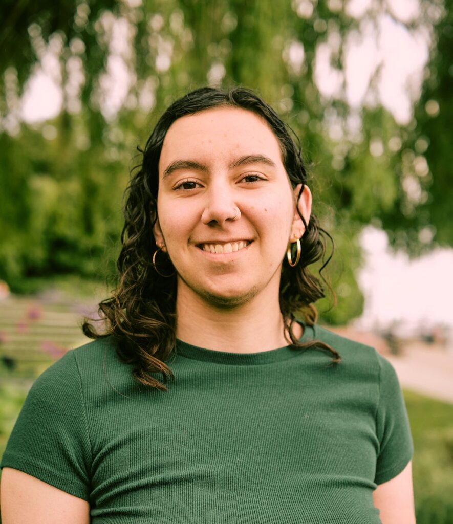 Jadi Darawi has dark curly hair, dark eyes, light skin, and gold hoop earrings and nose ring. They are wearing a dark green shirt and smiling at the camera. The background is a blurry green and gray garden scene.