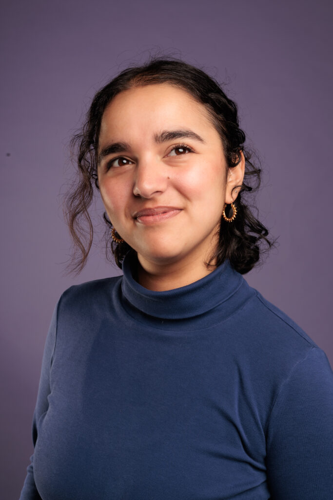 Summer Mahmud, a Pakistani-Tanzanian person with wavy dark brown hair smiles looking slightly up and off to the side. They wear a dark blue turtle-neck and small gold hooped earrings, in front of a lavender background.