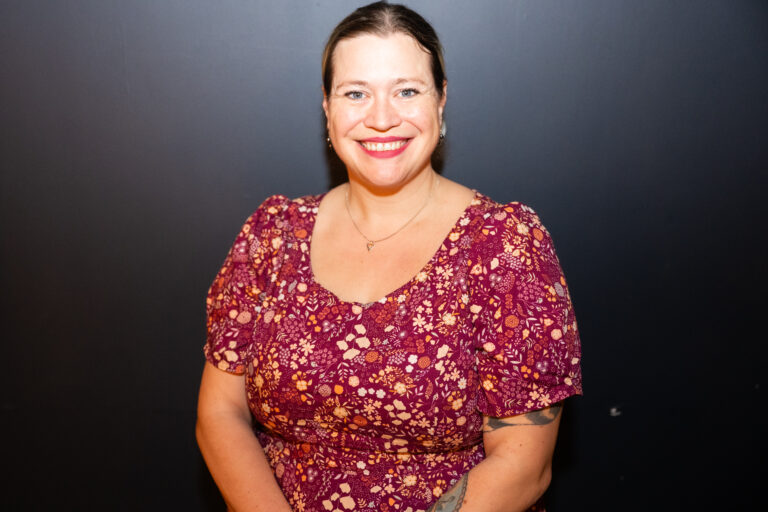 Photo of a white woman in her 30s. She is smiling to the camera in a red floral dress with matching red lipstick.