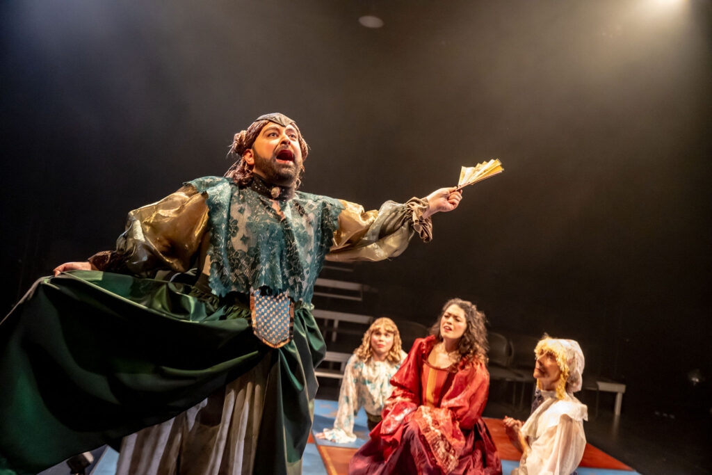 3 actors sit confused onstage looking up towards Izad Etemadi giving an impassioned speech.