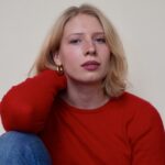photo of a white person in 20s. they have short blonde hair and a wearing a red sweater and jeans.