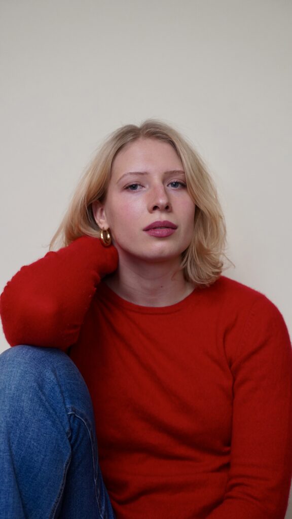 photo of a white person in 20s. they have short blonde hair and a wearing a red sweater and jeans.