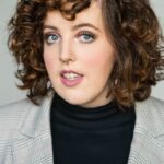 photo of a white person in their 20s. They have curly brown hair and are wearing a black turtle neck and gray blazer.