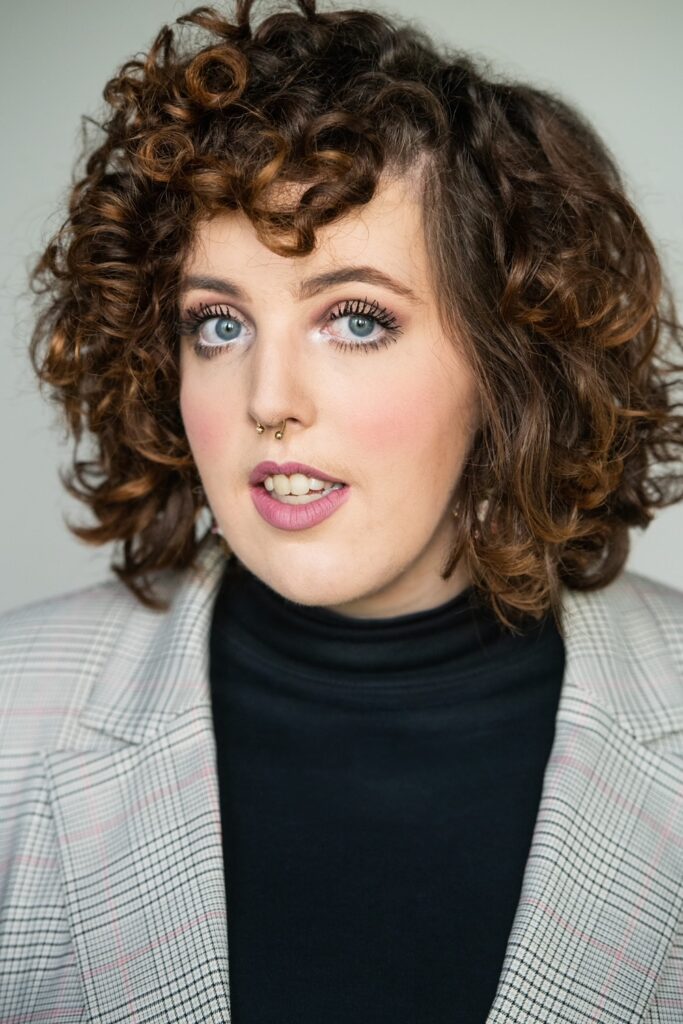 photo of a white person in their 20s. They have curly brown hair and are wearing a black turtle neck and gray blazer.