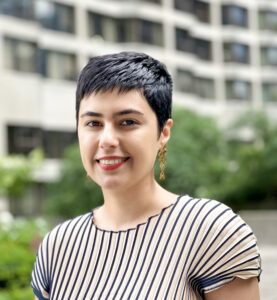 Headshot of a smiling person with short dark hair, wearing a cream and navy striped top and gold earrings, standing outdoors against a softly blurred urban background.