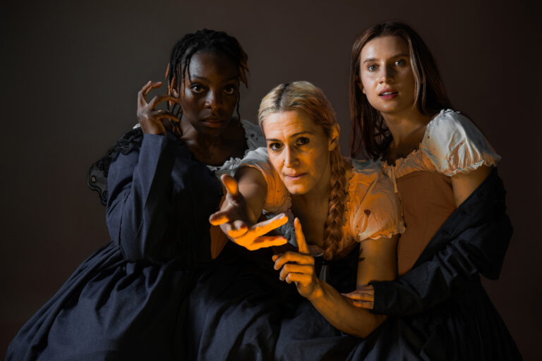 Three women in Victorian-style dresses lean toward the camera in a dark, theatrical portrait. The woman in the center reaches one hand forward, lit by a warm amber glow, while the women on either side stare ahead with tense, alert expressions. The background is shadowy and minimal, creating a moody Gothic atmosphere.