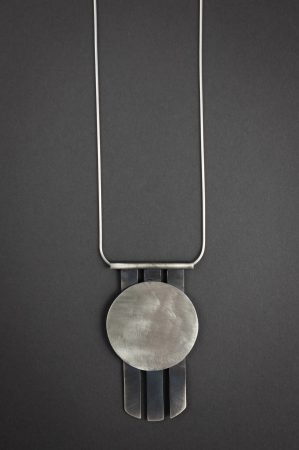 Against a grey background is a necklace that has a circular pendant with three metal spikes going vertically off of the bottom of the circle.