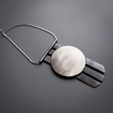 A necklace is lying diagonally on a grey surface. The necklace has a circular pendant with three metal spikes going vertically off of the bottom of the circle.