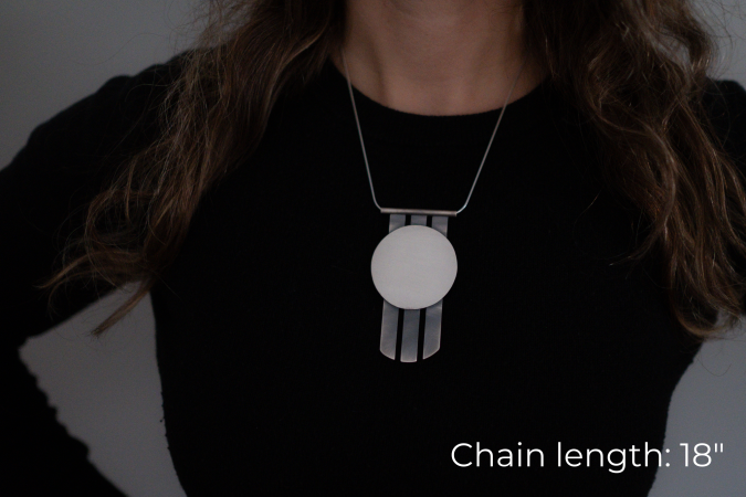 Miranda Britton, the artist, a white woman with brown hair wearing a black shirt, models a necklace that has a circular pendant with three metal spikes going vertically off of the bottom of the circle.