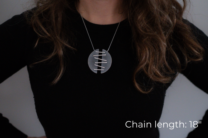 Miranda Britton, the artist, a white woman with brown hair wearing a black shirt, models a necklace that has two half-circle pendants that are connected with metal that is meant to look like stitches, holding the two halves together.