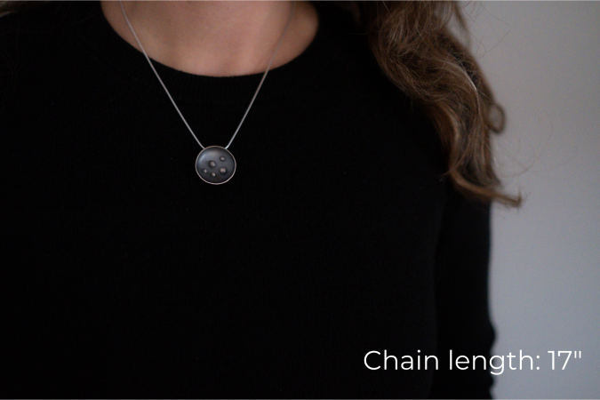 Miranda Britton, a white woman with brown hair wearing a black shirt, models the necklace that she has made. The necklace has a circular pendant with 5 dots in the centre, and is on a grey chain.