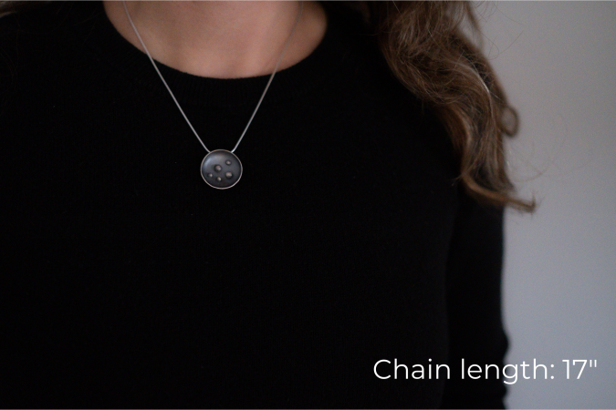 Miranda Britton, a white woman with brown hair wearing a black shirt, models the necklace that she has made. The necklace has a circular pendant with 5 dots in the centre, and is on a grey chain.