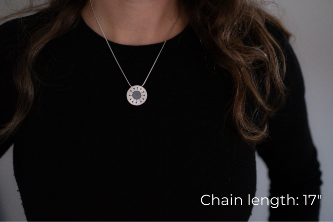 Miranda Britton, the artist, a white woman with brown hair wearing a black shirt, models a necklace that has a circular with a hole in the center, and 12 branches to much smaller holes coming off of the center.