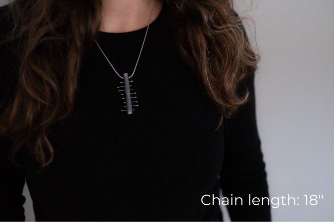 Miranda Britton, the artist, a white woman with brown hair wearing a black shirt, models a necklace that has a rectangle pendant with 8 smaller pieces of metal crossing the center piece.