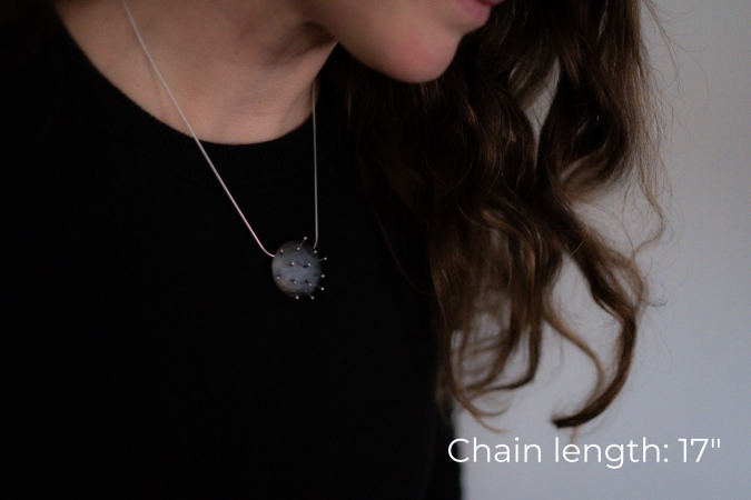 Miranda Britton, the artist, a white woman with brown hair wearing a black shirt, models a necklace that has a circular metal pendant that looks like a pin cushion, with 14 pins sticking out of the pendant.