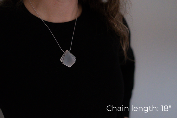 Miranda Britton, the artist, a white woman with brown hair wearing a black shirt, models a necklace that has a diamond shaped pendant at the center with two small triangle shapes on either side of the main pendant.