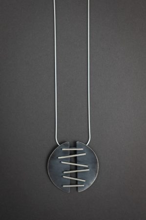 Against a grey background is a necklace that has two half-circle pendants that are connected with metal that is meant to look like stitches, holding the two halves together.