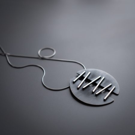 A necklace is lying diagonally on a grey surface. The necklace has two half-circle pendants that are connected with metal that is meant to look like stitches, holding the two halves together.