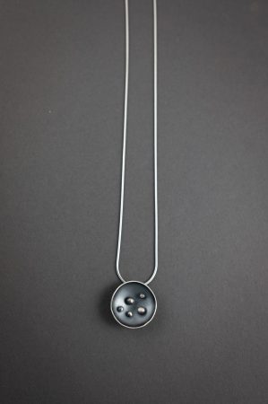A necklace against a grey background. The necklace has a circular pendant with 5 dots in the center, and is on a grey chain.