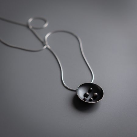 A necklace laying flat on a grey surface. The necklace has a circular pendant with 5 dots in the center, and is on a grey chain that is shown draped on an angle..