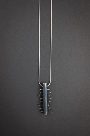 Against a grey background is a necklace that has a rectangle pendant with 8 smaller pieces of metal crossing the center piece.
