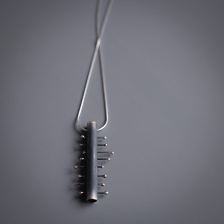A necklace is lying on a grey surface. The necklace has a rectangle pendant with 8 smaller pieces of metal crossing the center piece.
