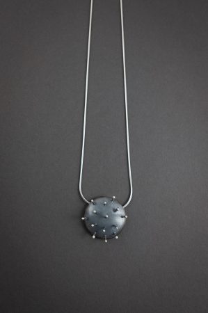 Against a grey background is a necklace that has a circular metal pendant that looks like a pin cushion, with 14 pins sticking out of the pendant.