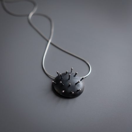 A necklace is lying diagonally on a grey surface. The necklace has a circular metal pendant that looks like a pin cushion, with 14 pins sticking out of the pendant.