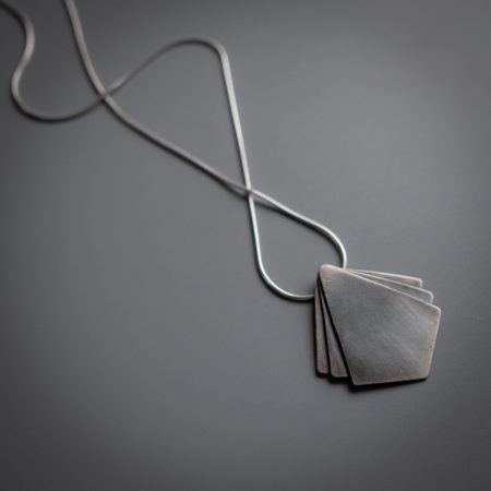A necklace is lying diagonally on a grey surface. The necklace has a diamond shaped pendant at the center with two small triangle shapes on either side of the main pendant.
